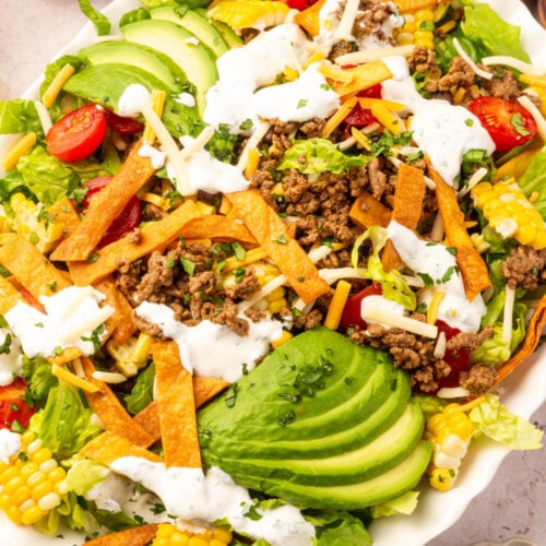 Gluten-free taco salad displayed on a table with fresh cilantro, wooden serving spoons, and a bowl of homemade tortilla strips.
