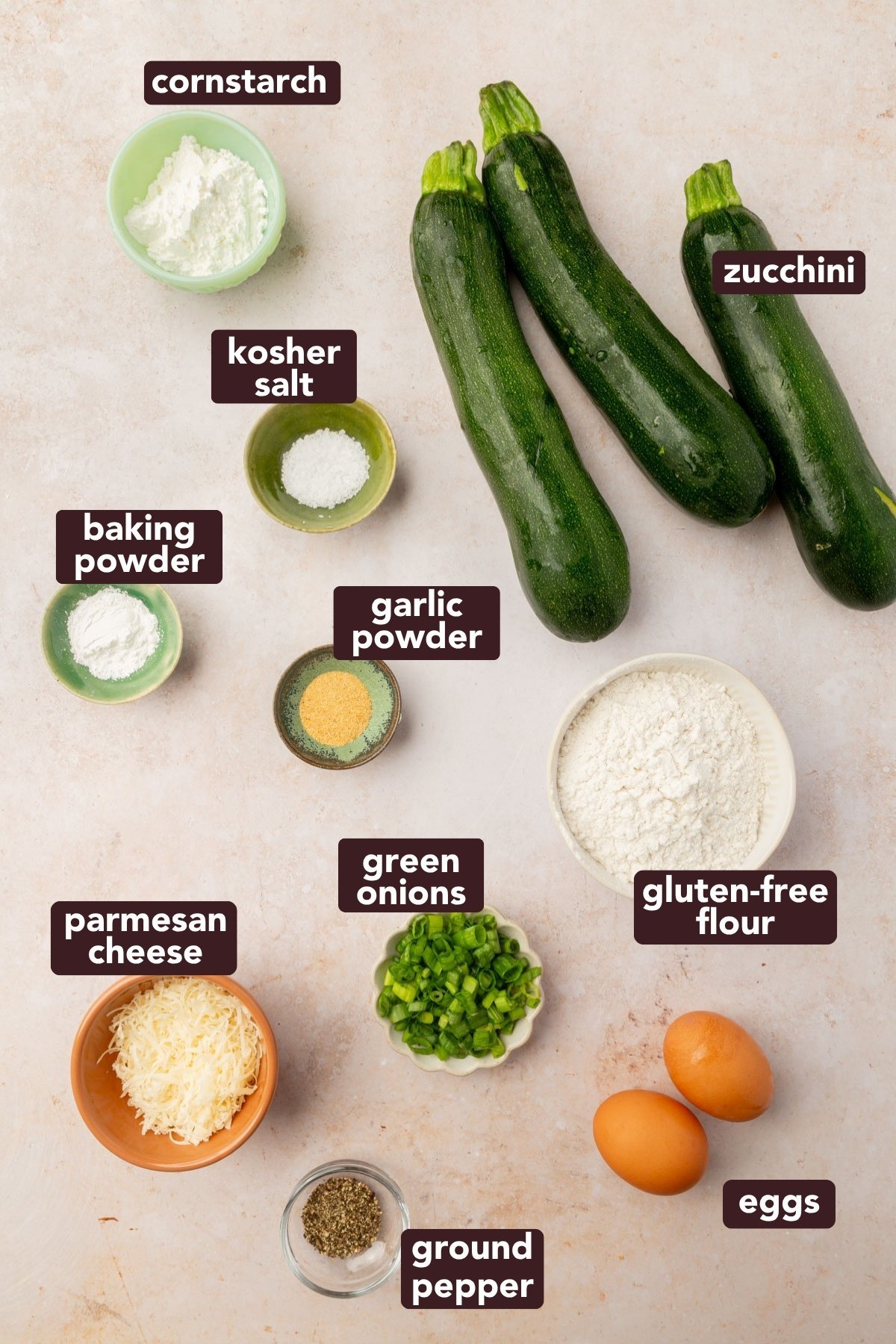Overhead view of ingredients for gluten-free zucchini fritters, including fresh zucchini, gluten-free flour, green onions, eggs, parmesan, and spices.