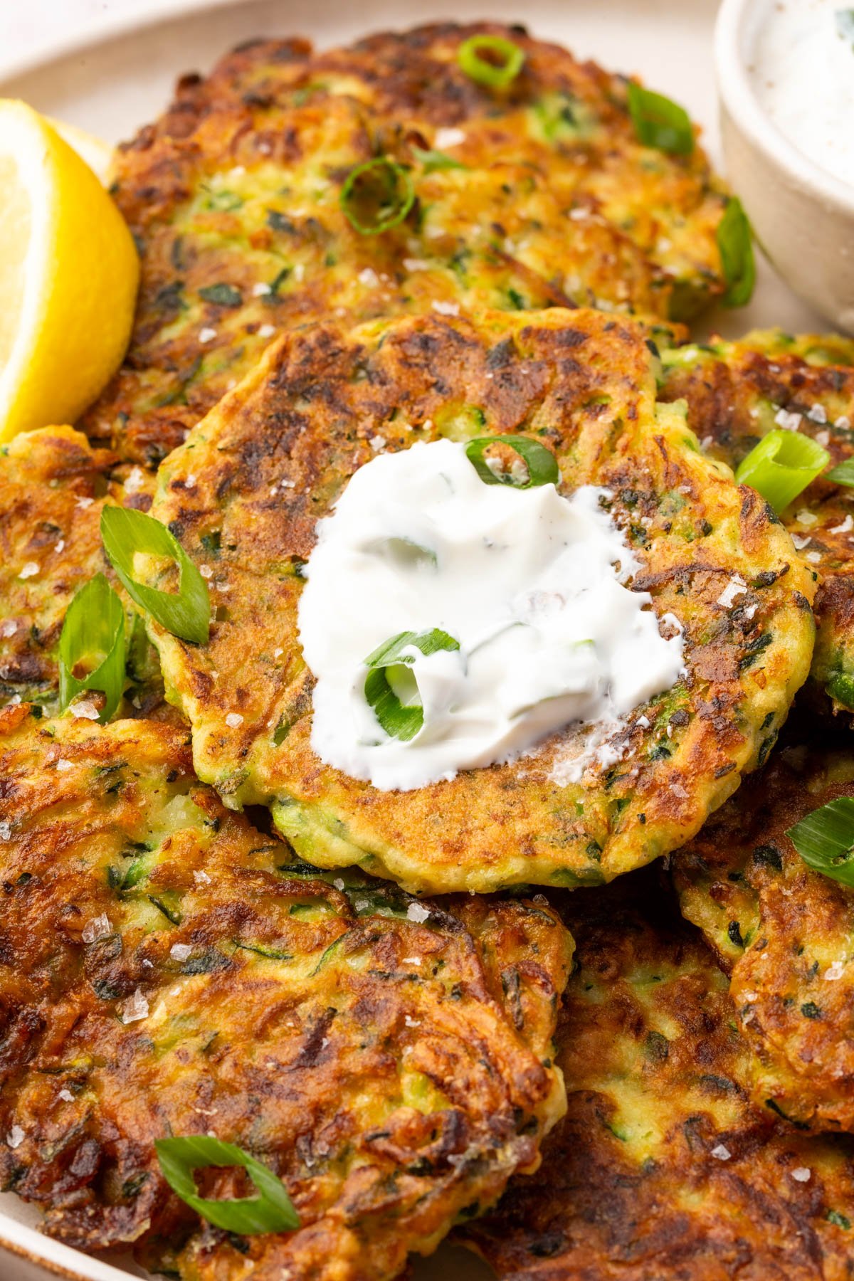 Close-up of a golden-brown zucchini fritter with crispy edges, topped with sour cream and fresh green onions.