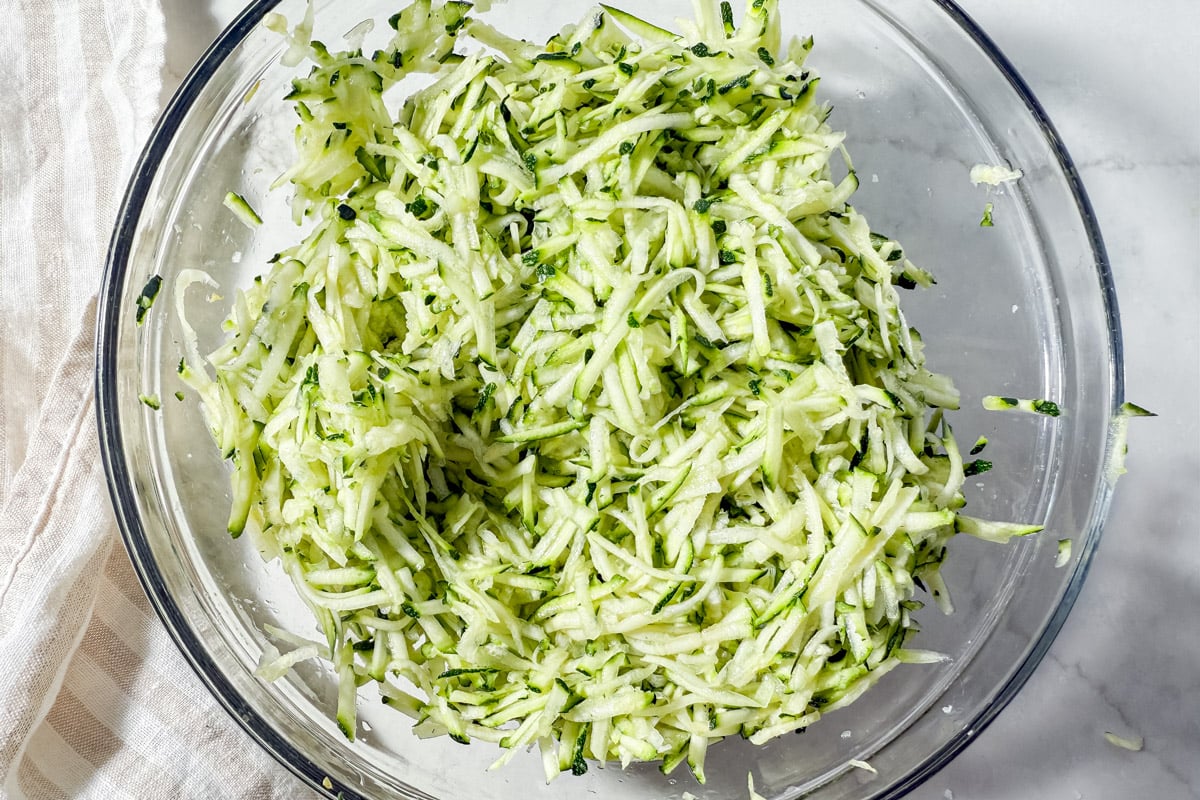 Freshly shredded zucchini in a clear glass mixing bowl on a white countertop.