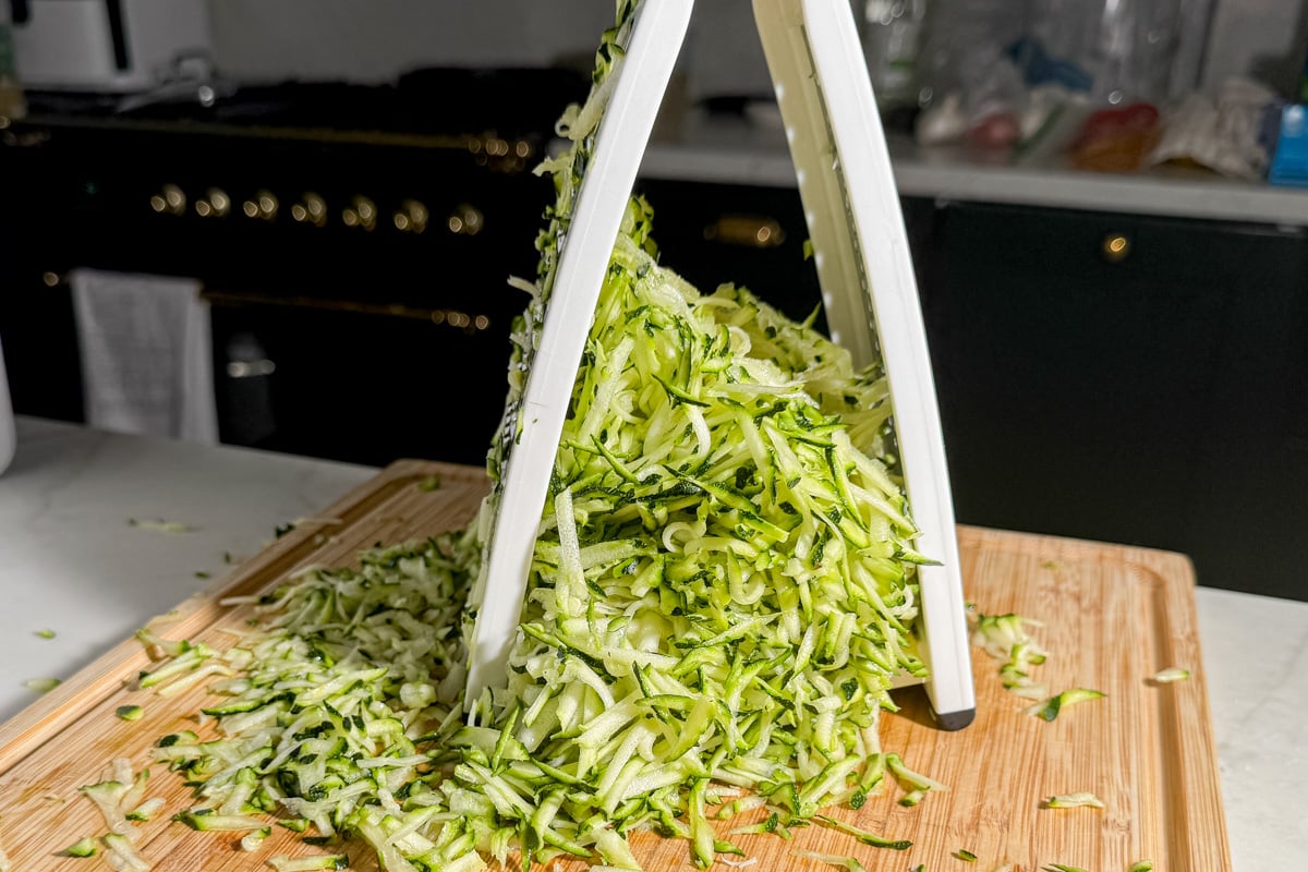 Zucchini being shredded with a box grater over a wooden cutting board, creating a large pile of ribbons.