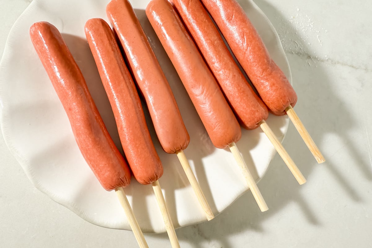 Six hot dogs on wooden skewers arranged neatly on a white plate, ready for battering.