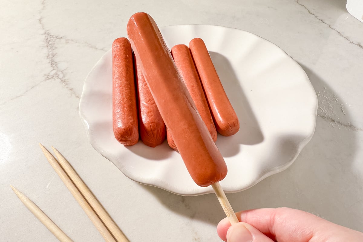 A hand holding a hot dog on a wooden skewer over a plate of unskewered hot dogs.