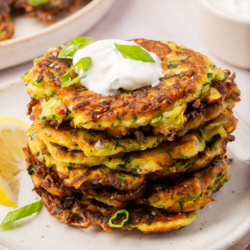 Stack of crispy gluten-free zucchini fritters served on a white plate, topped with sour cream and sliced green onions.