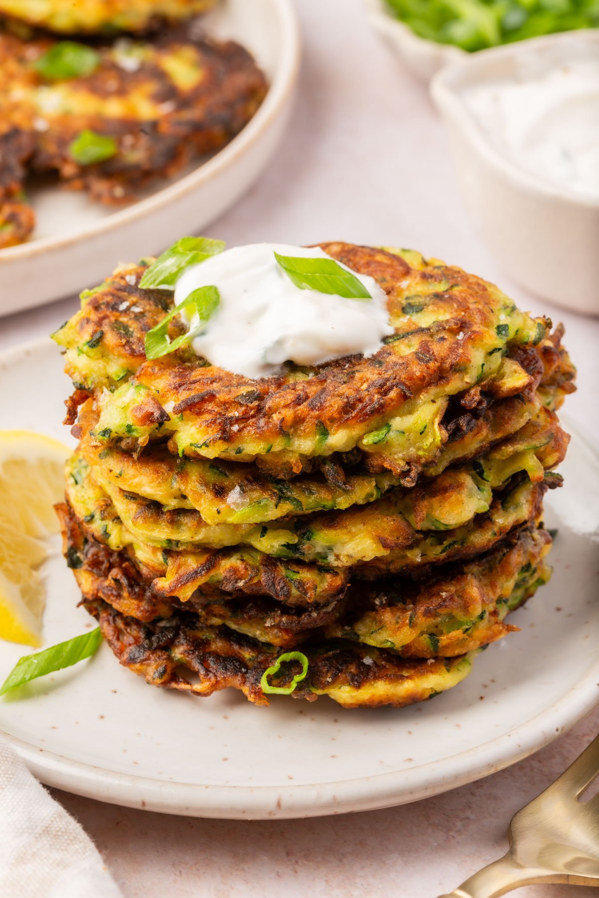 Stack of crispy gluten-free zucchini fritters served on a white plate, topped with sour cream and sliced green onions.