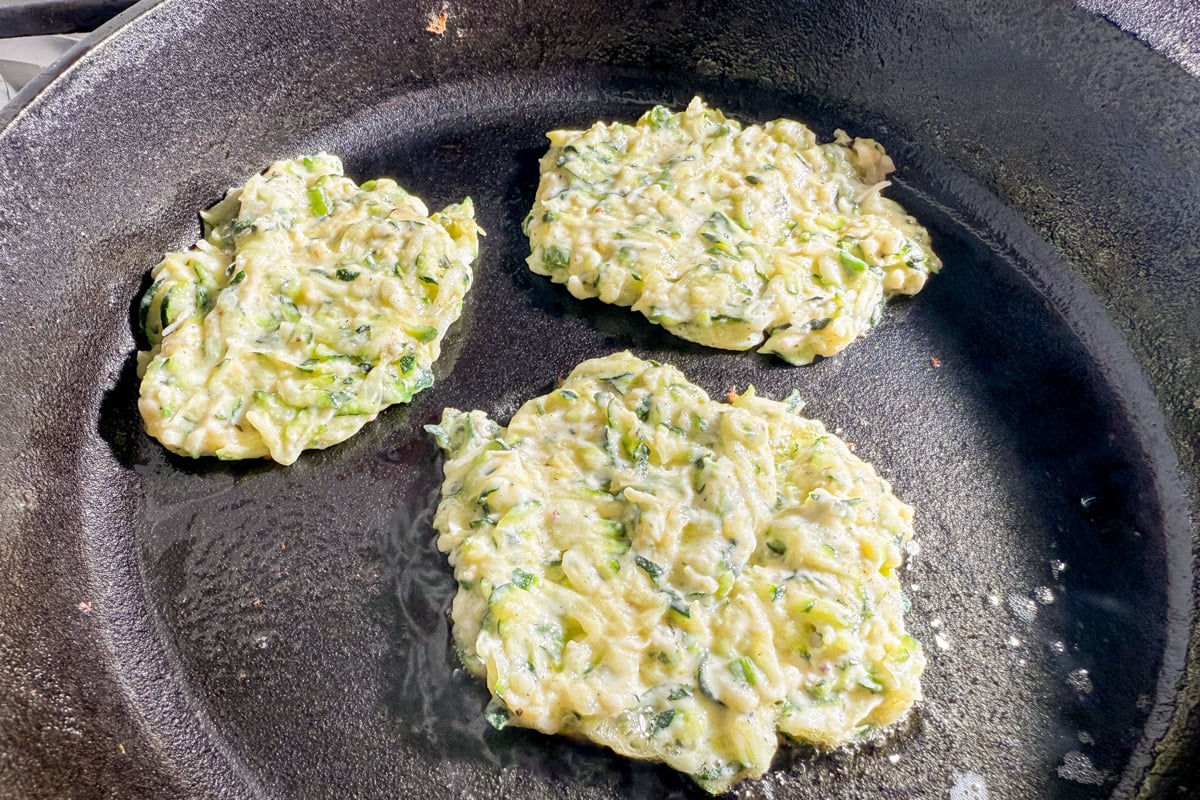 Four crispy zucchini fritters frying in a cast iron skillet with bubbling oil.