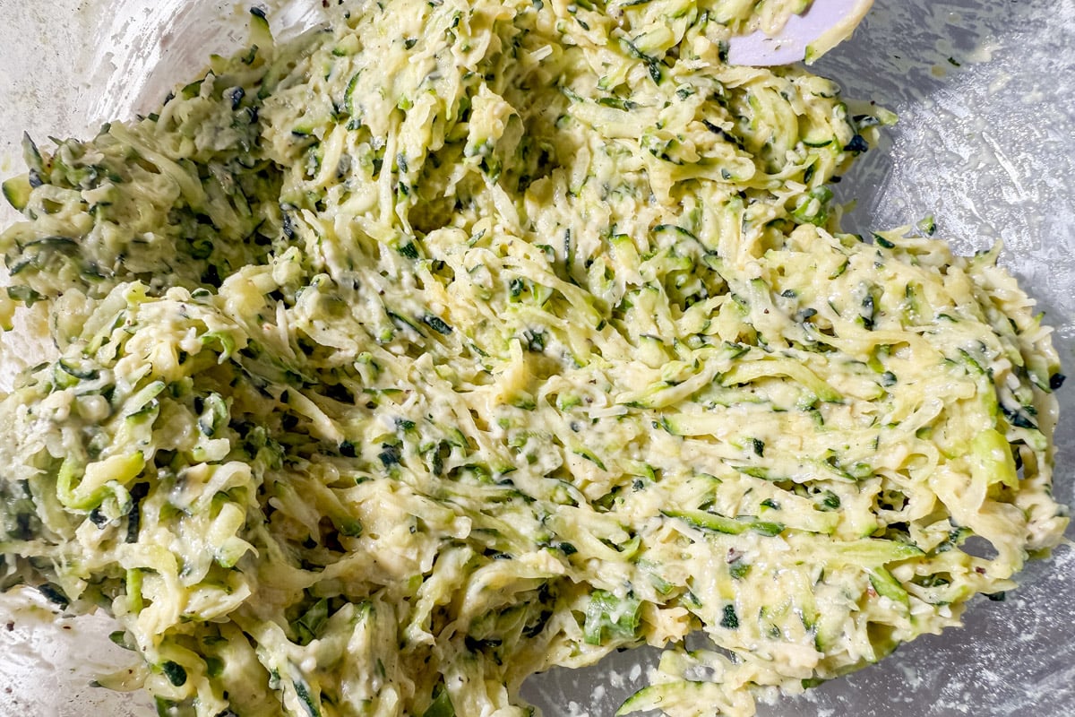 Mixed zucchini fritter batter in a glass bowl with visible shredded zucchini and flour.
