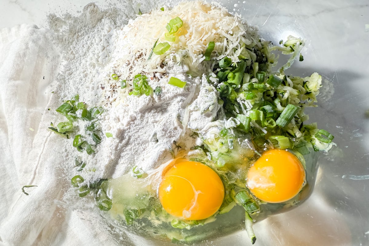 Overhead view of zucchini fritter ingredients in a glass bowl, including eggs, gluten-free flour, shredded zucchini, parmesan, and green onion.