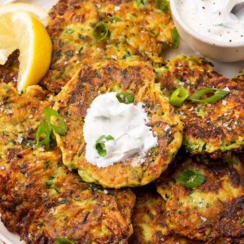 Gluten-free zucchini fritters topped with a dollop of sour cream and garnished with green onion, served with lemon slices and creamy dip on the side.