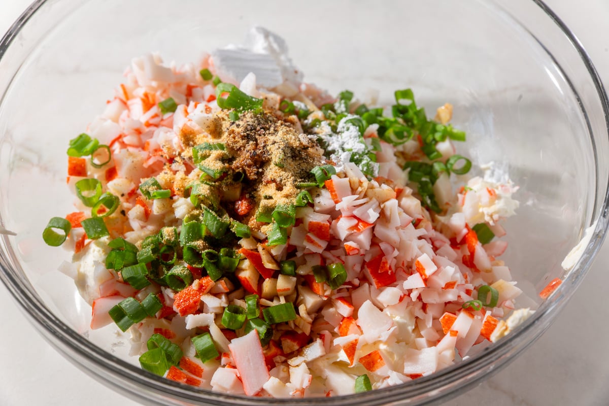 Glass bowl filled with chopped crab, green onions, cream cheese, and seasonings before mixing into crab rangoon filling.