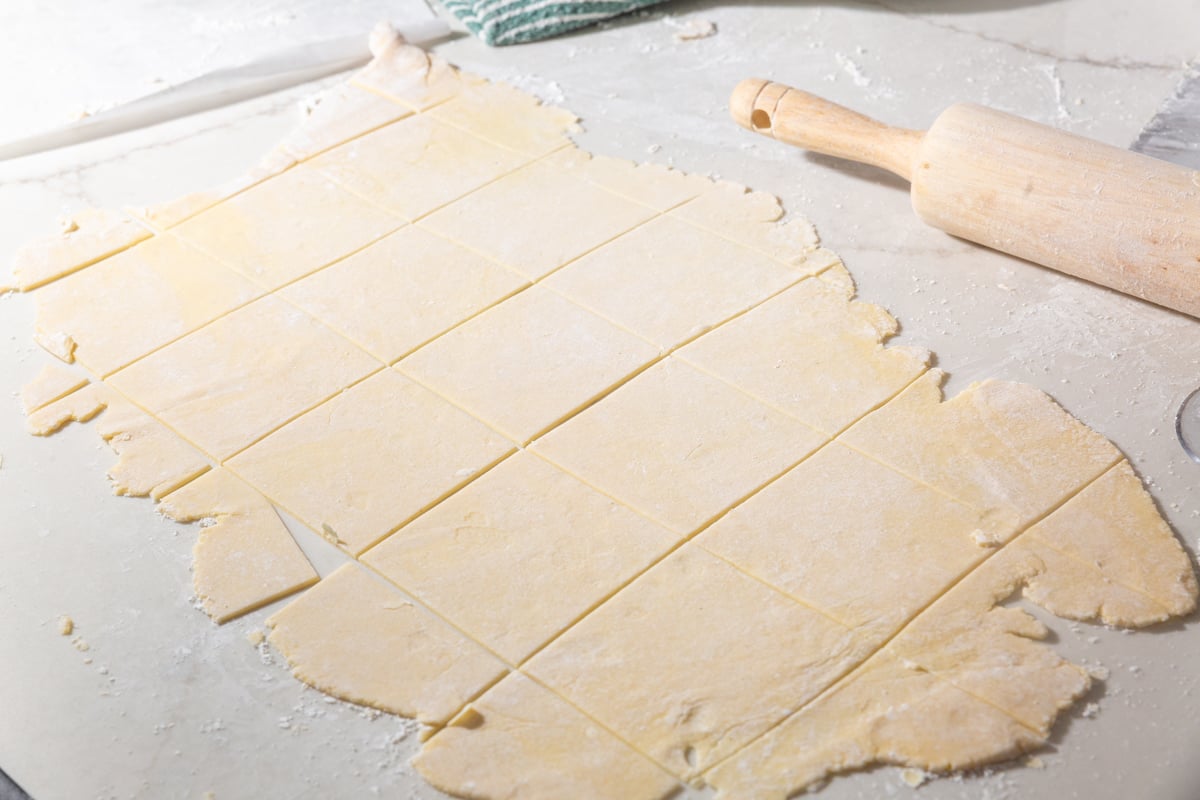 Rolled out gluten-free wonton dough cut into squares on a floured surface with a rolling pin nearby.