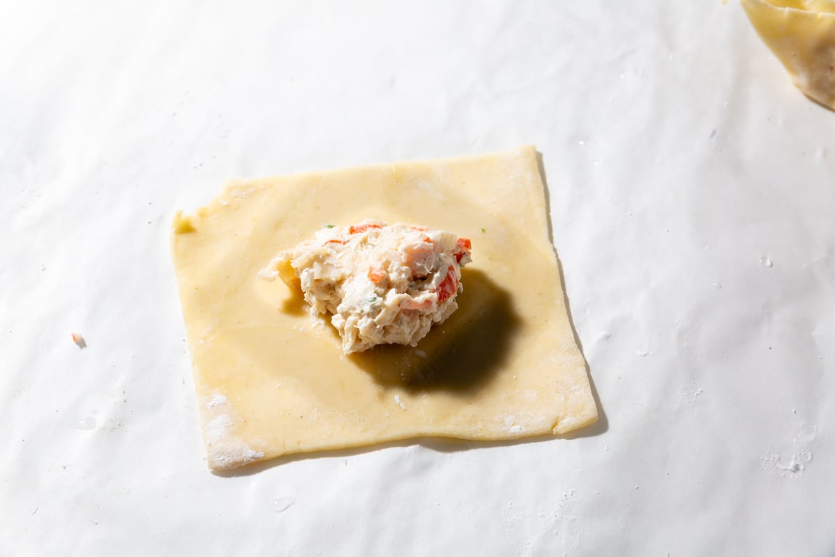 A square gluten-free wonton wrapper topped with a scoop of creamy crab rangoon filling and a diamond of water surrounding the filling.