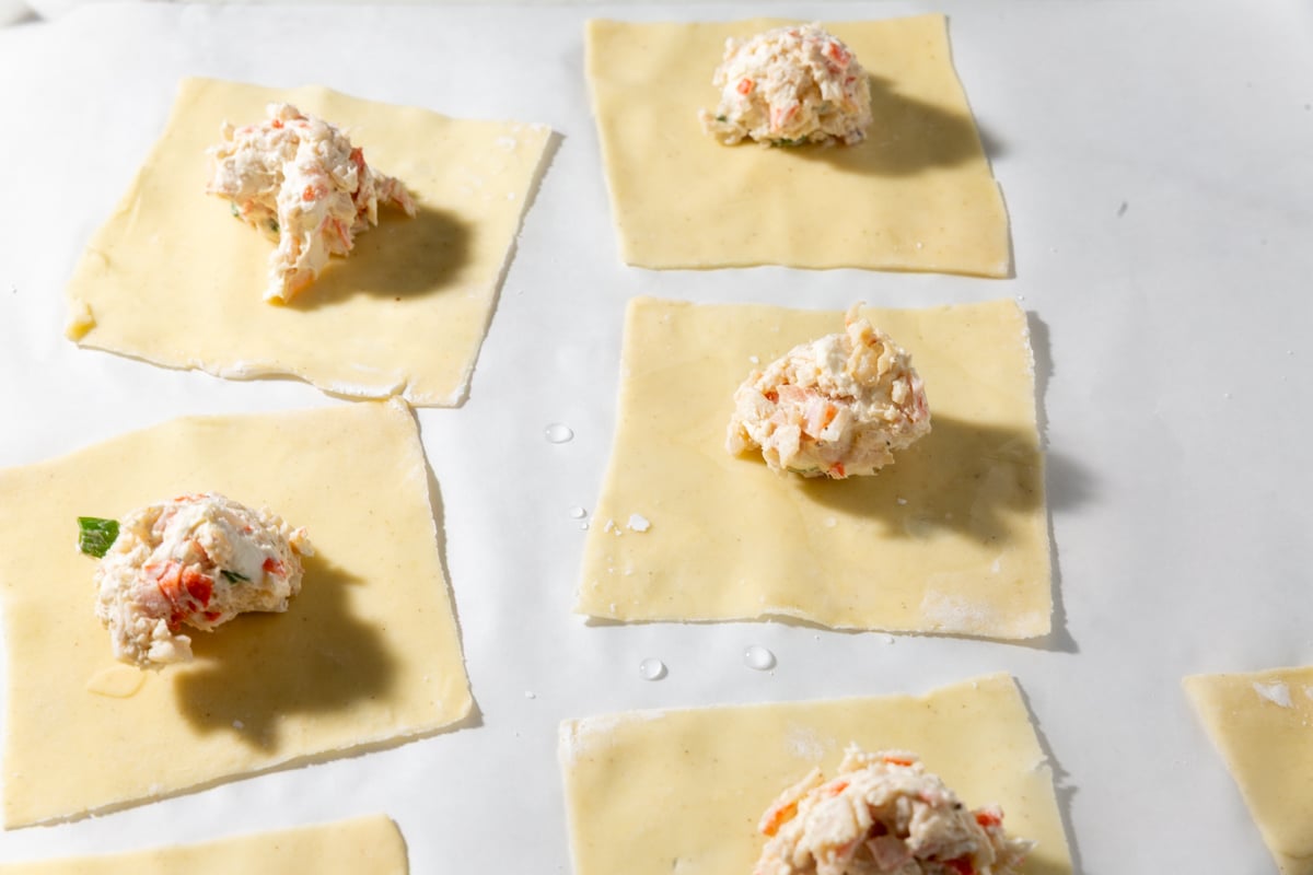 Squares of gluten-free wonton wrappers topped with spoonfuls of creamy crab rangoon filling, ready to be folded.
