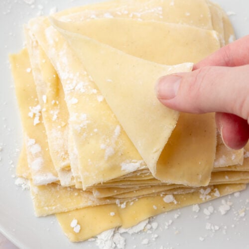 Hand peeling apart thin sheets of homemade gluten-free wonton wrappers on a white plate.