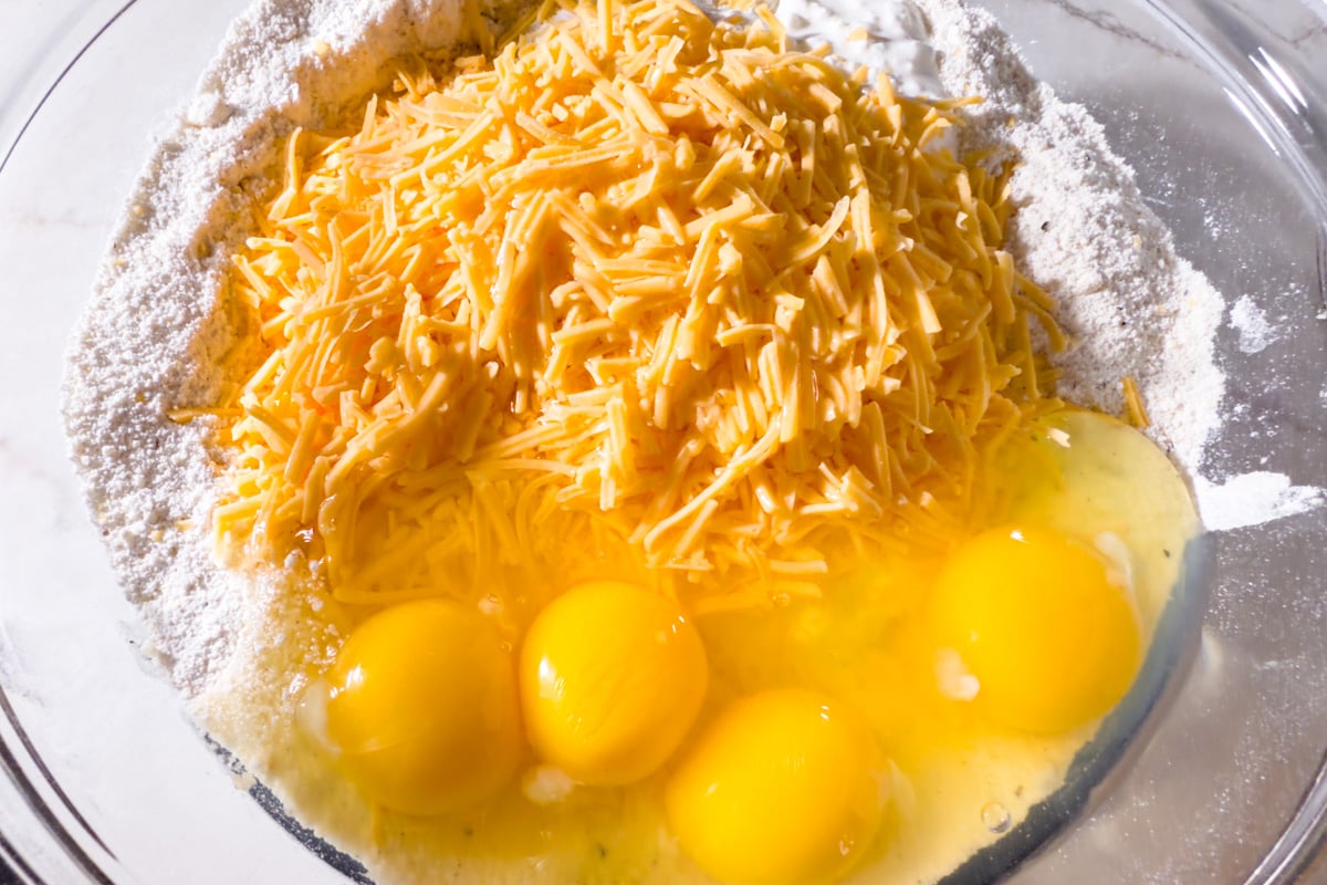 Shredded cheddar cheese and cracked eggs added on top of the dry ingredients in a glass mixing bowl.