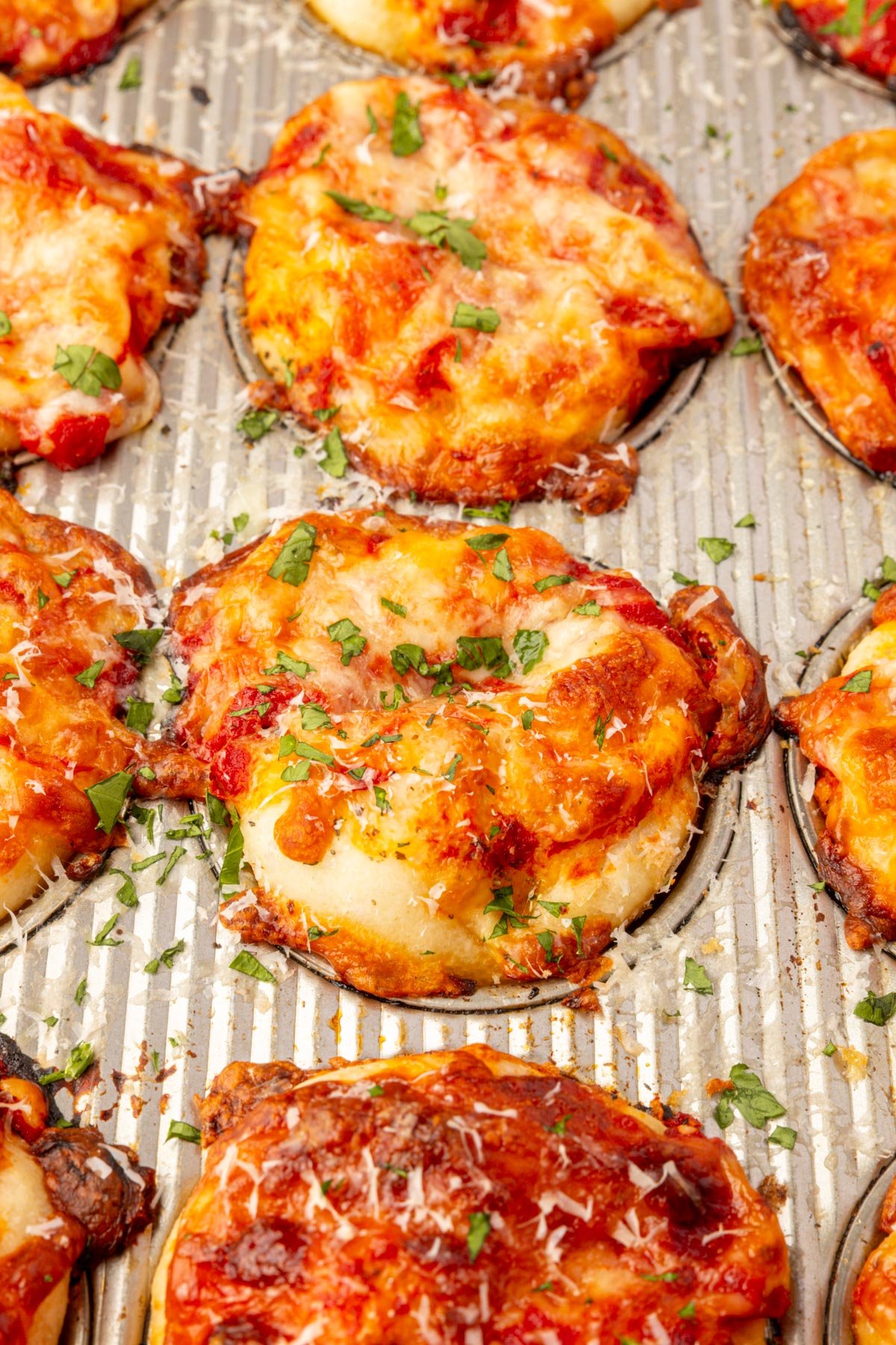 Baked gluten-free focaccia pizza muffins in a muffin tin topped with melted mozzarella, marinara, and herbs.