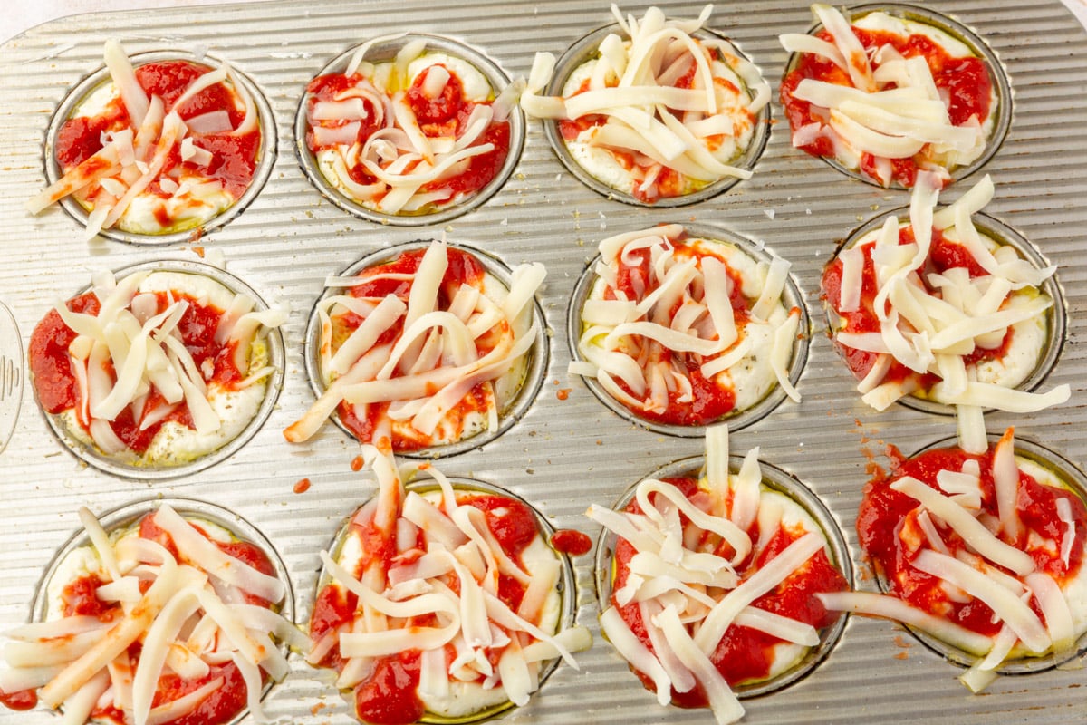 Muffin tin filled with focaccia dough topped with pizza sauce and shredded mozzarella cheese before baking.