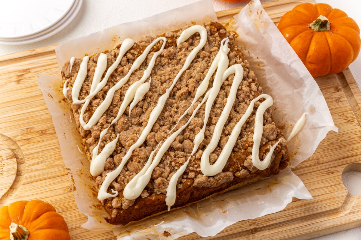 Freshly baked gluten-free pumpkin coffee cake with crumb topping and cream cheese glaze drizzle on parchment paper, surrounded by small pumpkins.