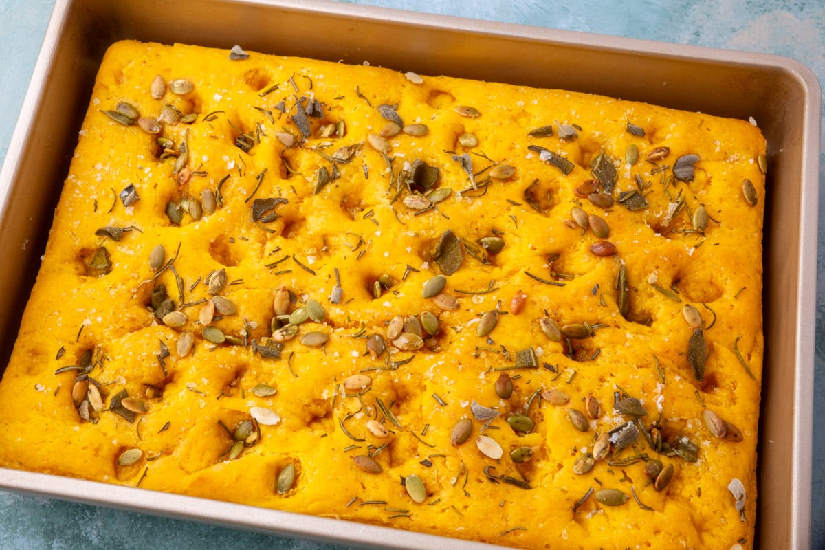 Freshly baked gluten-free pumpkin focaccia in a pan topped with rosemary, sage, pepitas, and flaky sea salt.