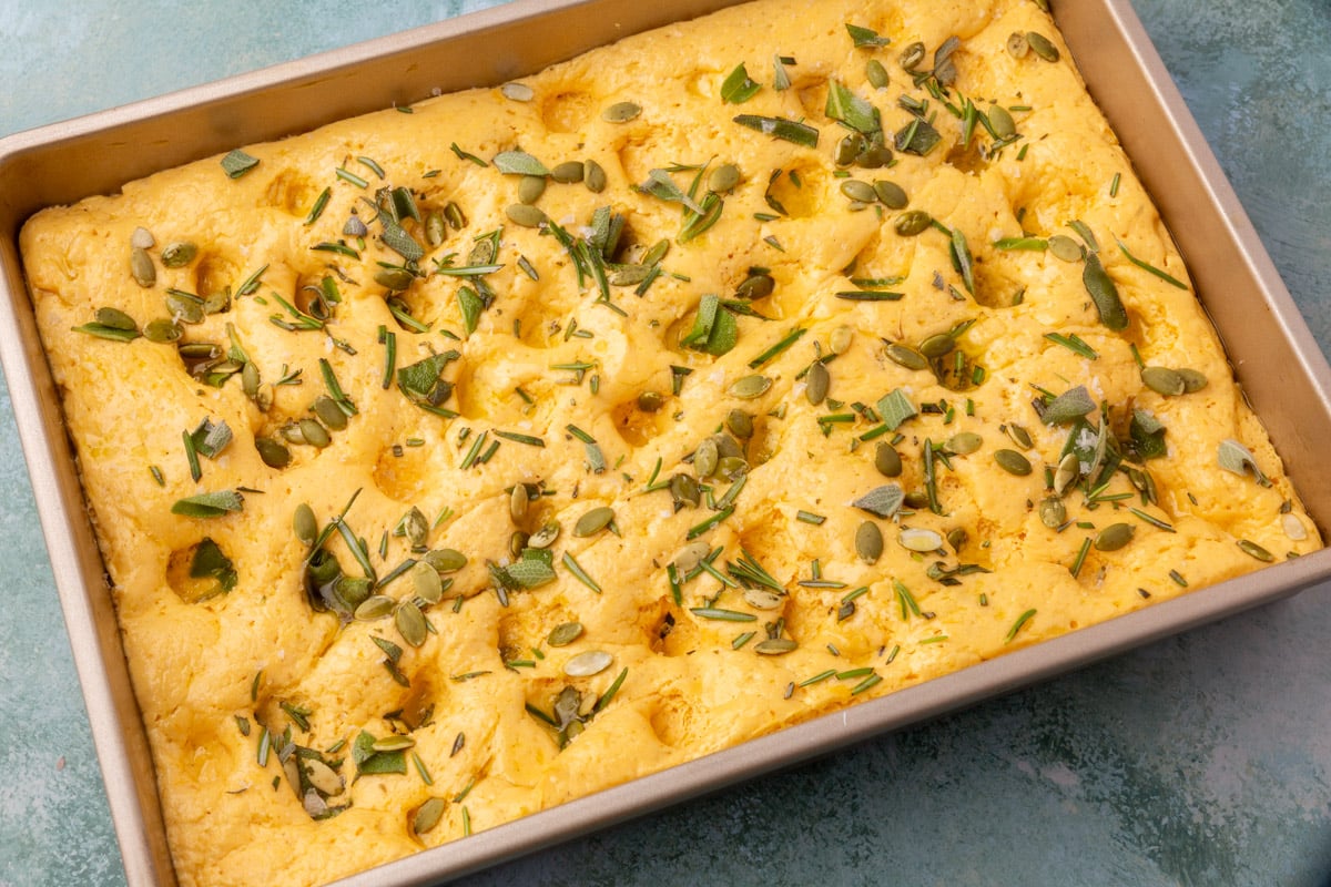 Unbaked pumpkin focaccia dough dimpled and topped with rosemary, sage, pepitas, and sea salt.