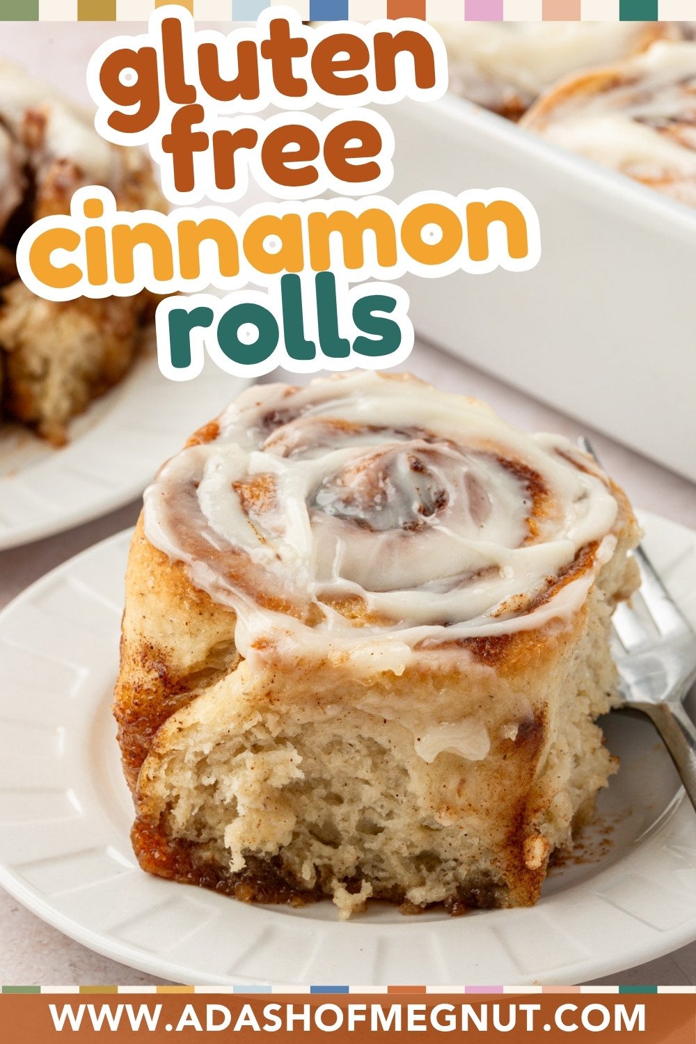 Close-up of a frosted gluten-free cinnamon roll on a plate with a pan of rolls in the background.