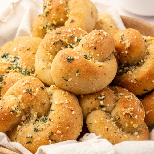 A wooden bowl filled with gluten-free garlic knots topped with garlic and herbs, with a small dish of marinara sauce in the background.