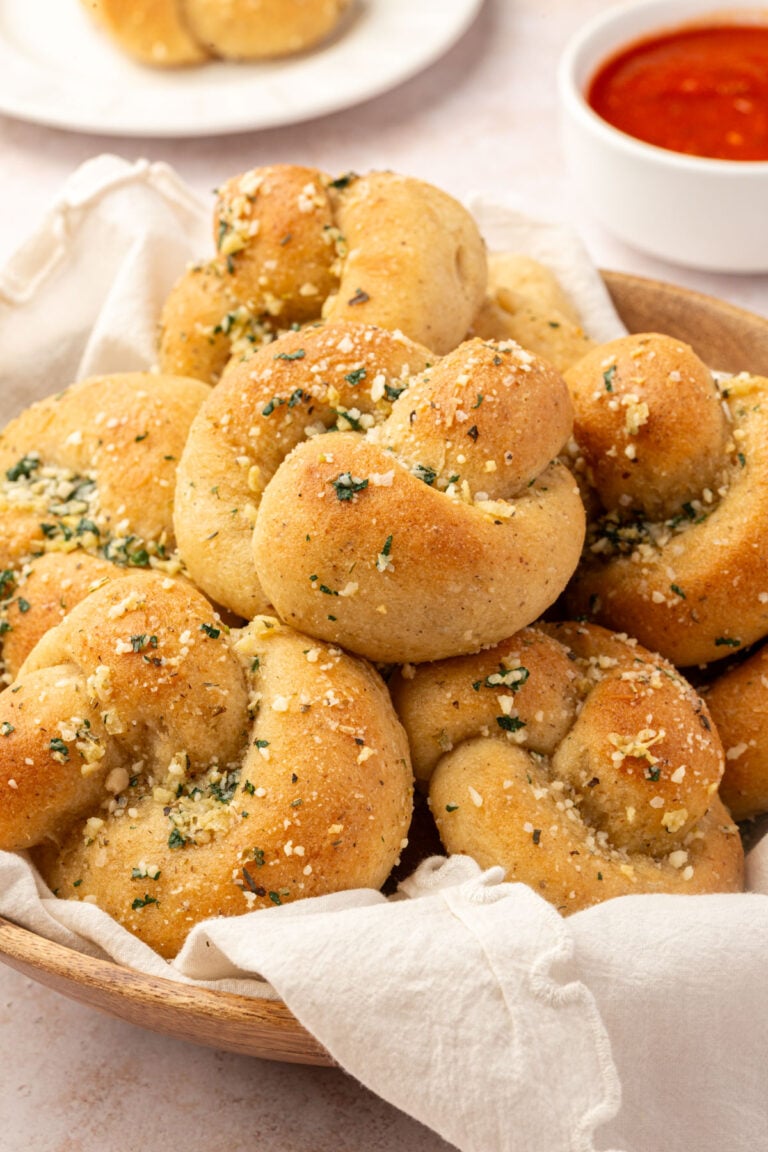 A wooden bowl filled with gluten-free garlic knots topped with garlic and herbs, with a small dish of marinara sauce in the background.