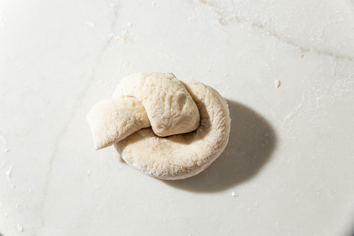 A shaped piece of dough twisted into a garlic knot on a floured surface before rising.