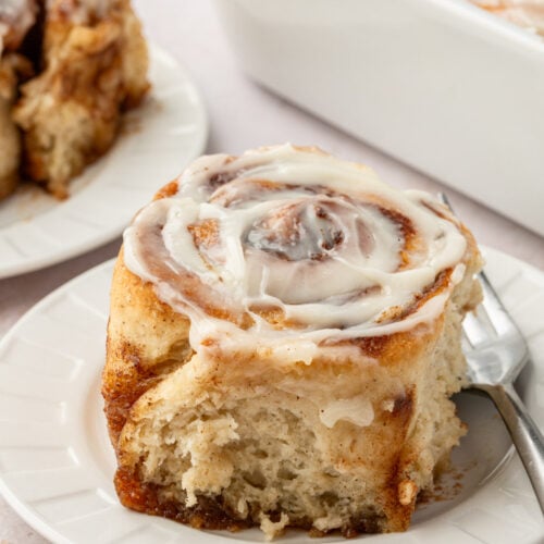 Gooey gluten-free cinnamon roll topped with cream cheese frosting, served on a white plate with a fork.
