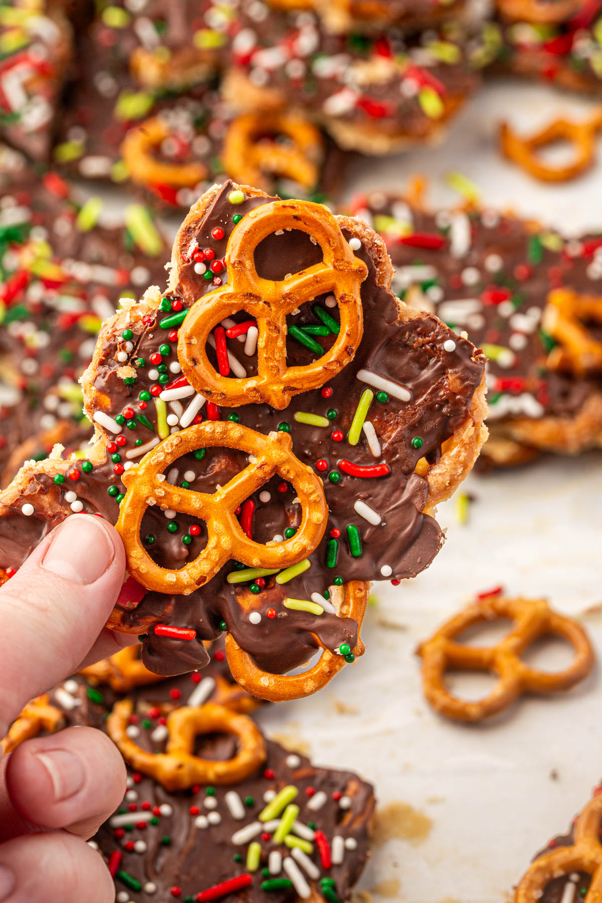 Hand holding piece of gluten-free Christmas pretzel bark with chocolate, caramel, colorful holiday sprinkles, and crushed pretzels.