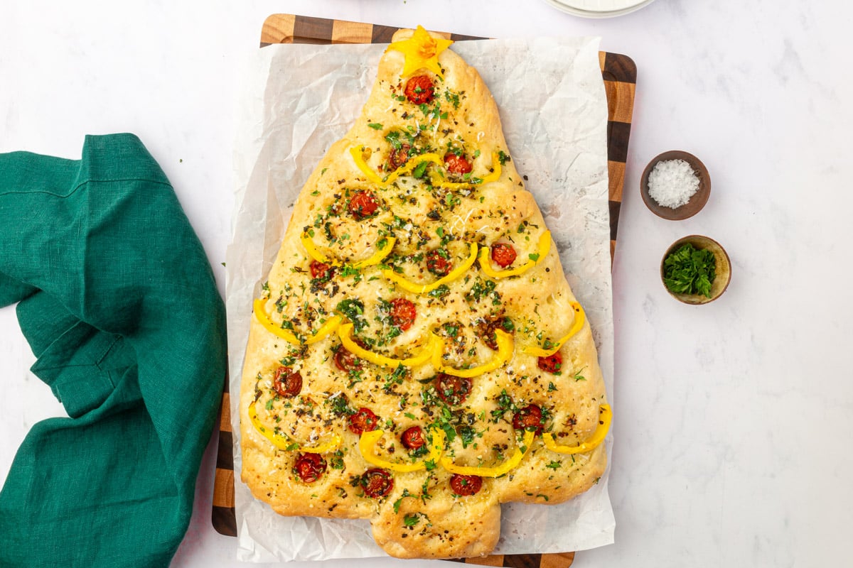 Golden baked Christmas tree focaccia on wood board with green napkin, parsley and flaky salt in small bowls.