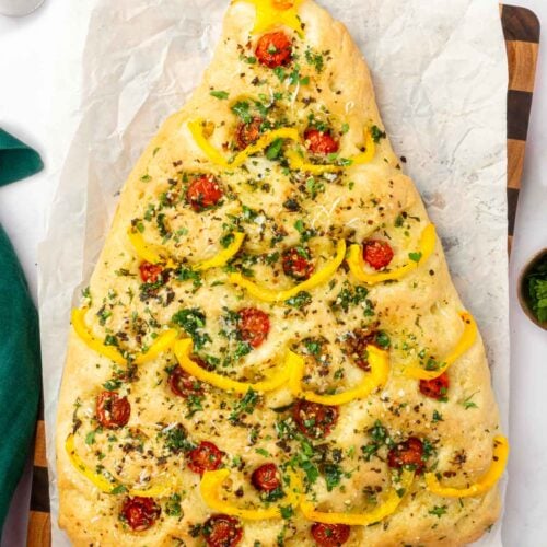 Overhead view of baked Christmas tree focaccia decorated with yellow bell peppers and cherry tomatoes on wood cutting board.