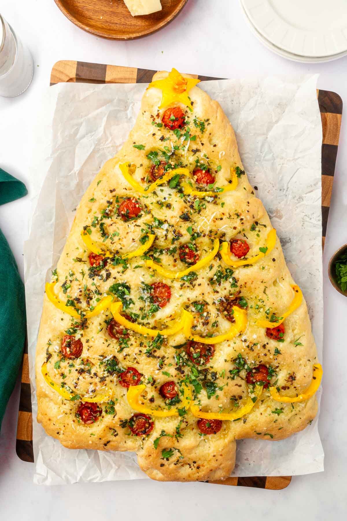 Overhead view of baked Christmas tree focaccia decorated with yellow bell peppers and cherry tomatoes on wood cutting board.