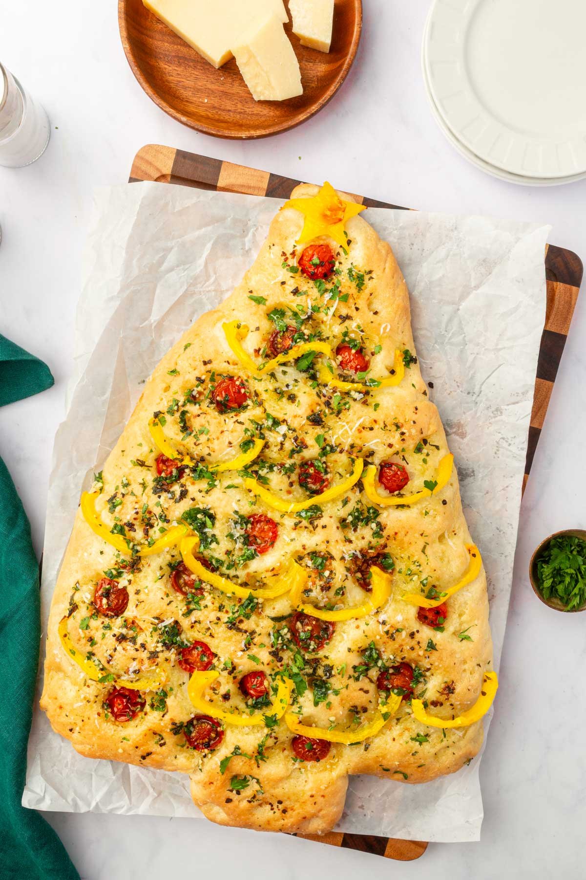 Gluten-free Christmas tree focaccia on parchment-lined wood board with yellow pepper star and garland, cherry tomato ornaments, and parmesan cheese.