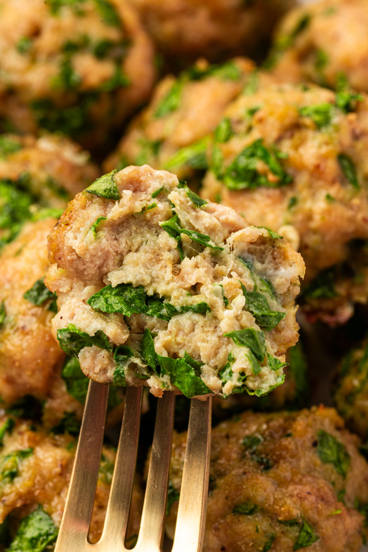 A close-up of a fork holding a meatball with visible chopped spinach inside, showing its juicy and tender texture. More spinach-filled meatballs are blurred in the background.