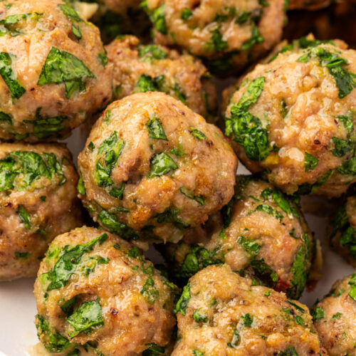 A close-up of several baked meatballs on a white plate, mixed with visible green herbs and a golden-brown exterior.