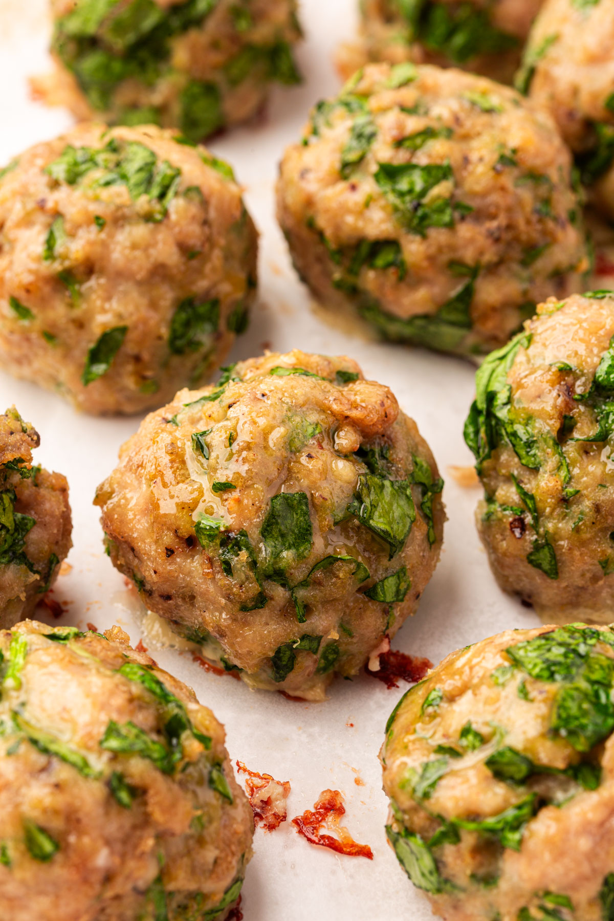 Close-up of freshly baked meatballs with visible chopped green spinach mixed in, resting on a sheet of parchment paper. The meatballs appear juicy and golden brown.