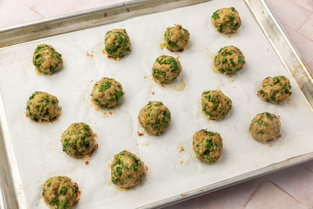 A baking sheet lined with parchment paper holds fifteen evenly spaced, cooked turkey meatballs freshly baked from the oven.