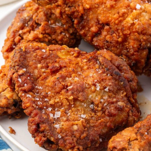 Close up of crispy gluten-free fried chicken with craggy coating and flaky sea salt on white platter.