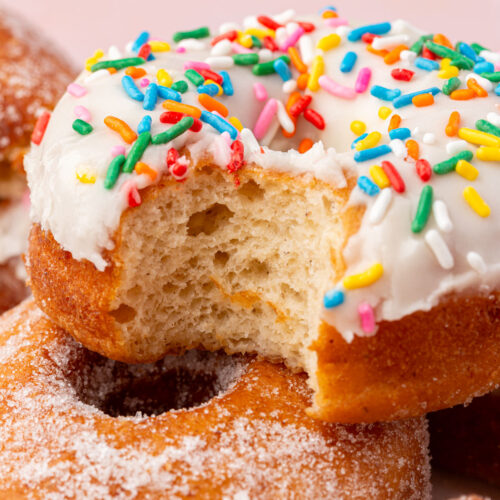 A close-up of a donut topped with white icing and colorful sprinkles, showing a bite taken out. The donut rests on others coated with granulated sugar, against a soft pink background.