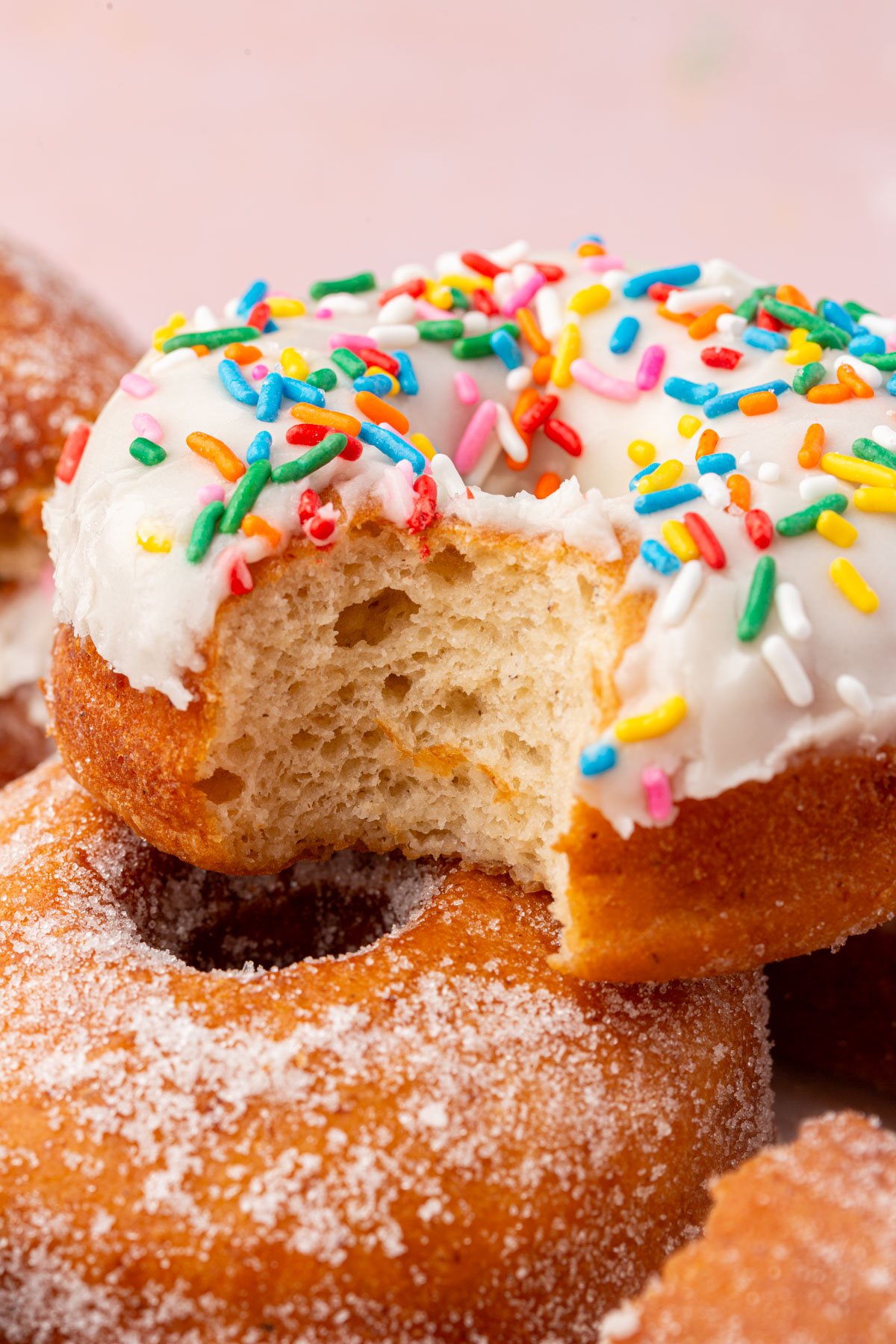 A close-up of a donut topped with white icing and colorful sprinkles, showing a bite taken out. The donut rests on others coated with granulated sugar, against a soft pink background.