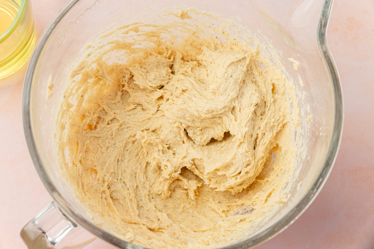 A glass mixing bowl filled with creamy, light brown cookie dough, partially mixed, sitting on a light pink surface. A glass cup with a yellow liquid is partially visible beside the bowl.