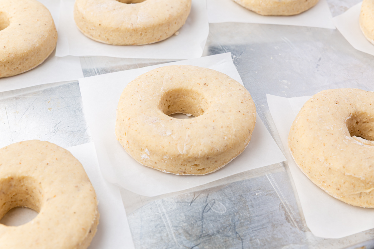 Unbaked, round doughnuts with holes in the center rest on individual squares of parchment paper, arranged on a metal surface.