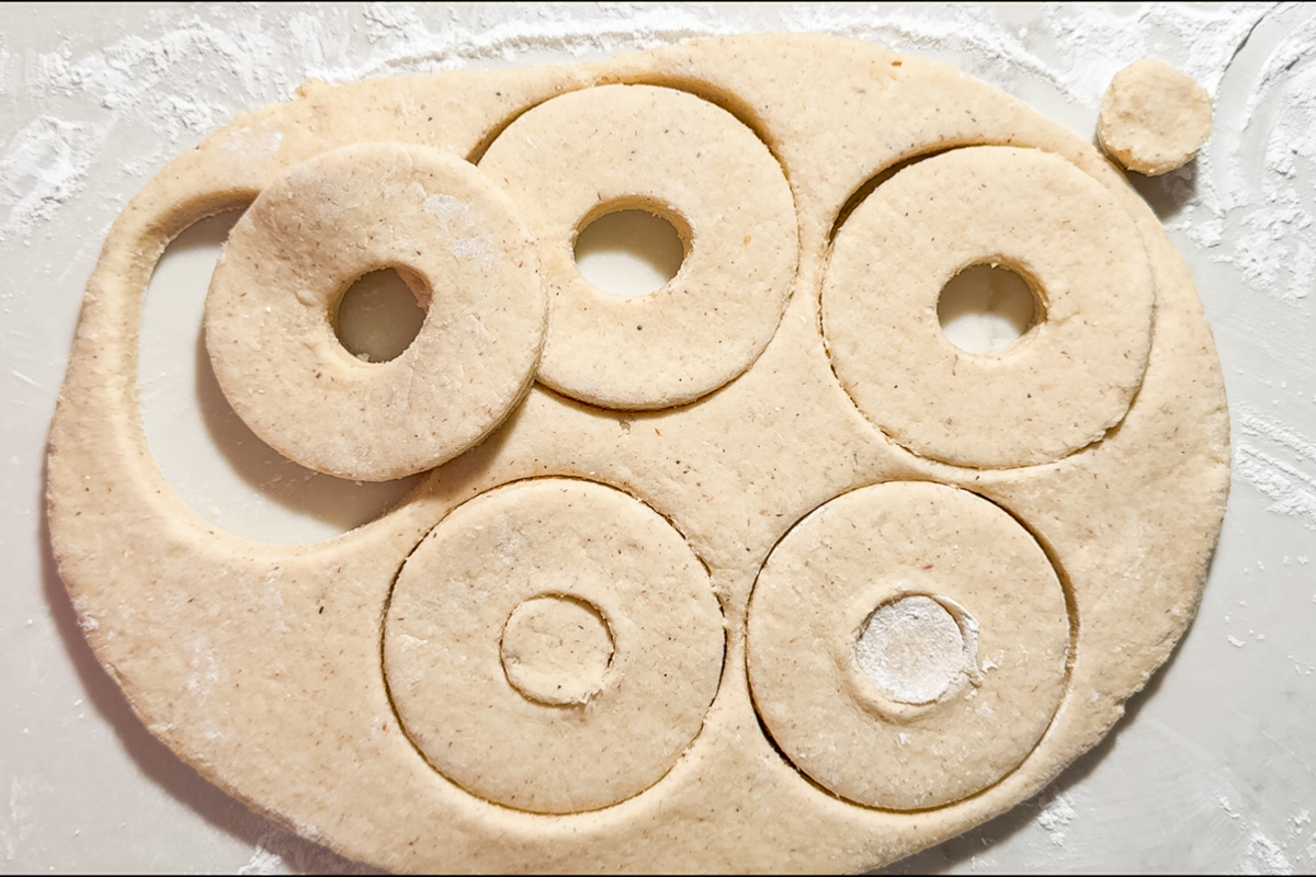 Gluten free fried donuts take shape as cookie dough is rolled out on a floured surface, with round shapes cut out and smaller circles removed from the center, creating classic donut forms.