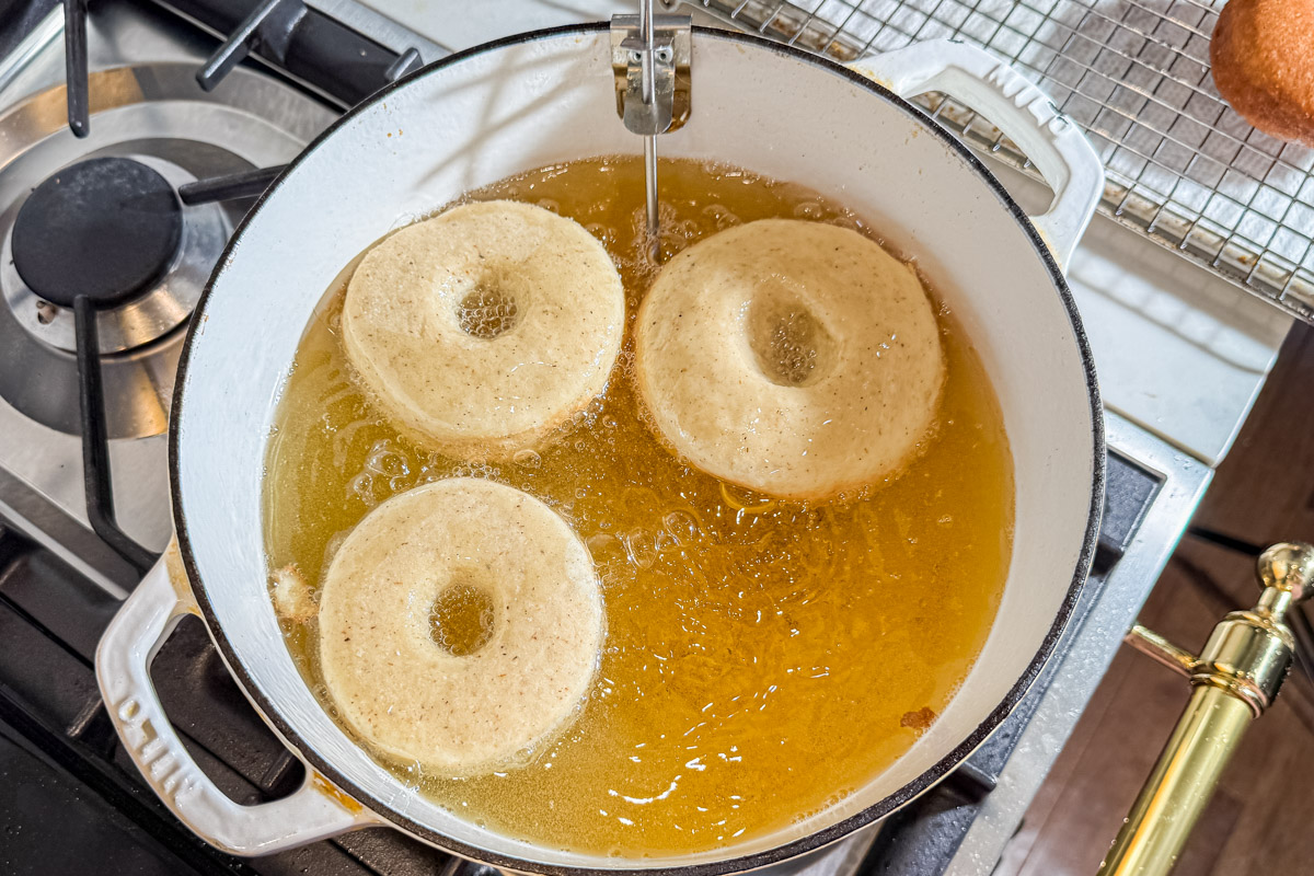Three unglazed donuts are frying in hot oil inside a white Dutch oven on a stovetop. A metal thermometer is clipped to the pot, and a cooling rack with a finished donut is visible nearby.