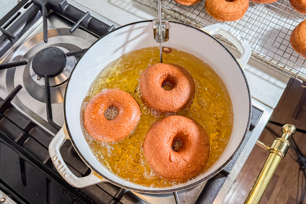 Three donuts are being fried in hot oil in a white pot on a stove, with more finished donuts draining on a cooling rack nearby.
