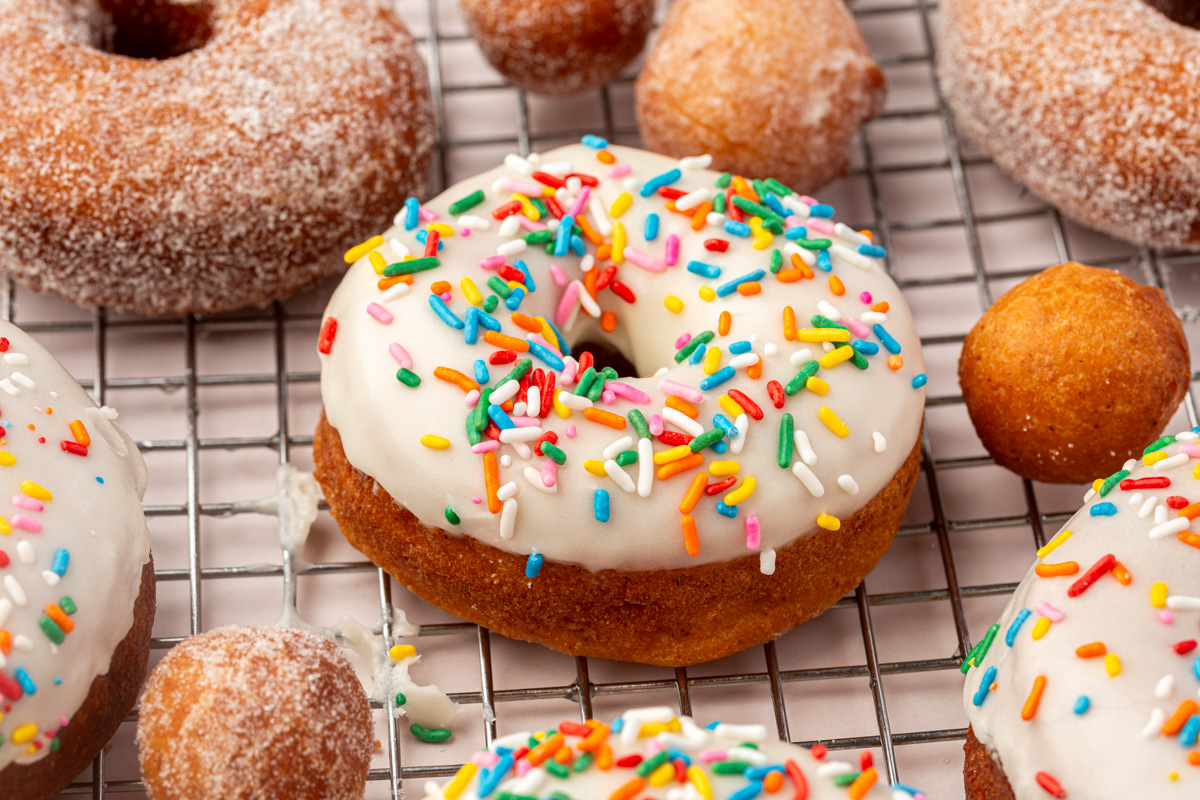 A wire cooling rack with donuts topped with white icing and colorful rainbow sprinkles, surrounded by plain sugared donuts and round donut holes.