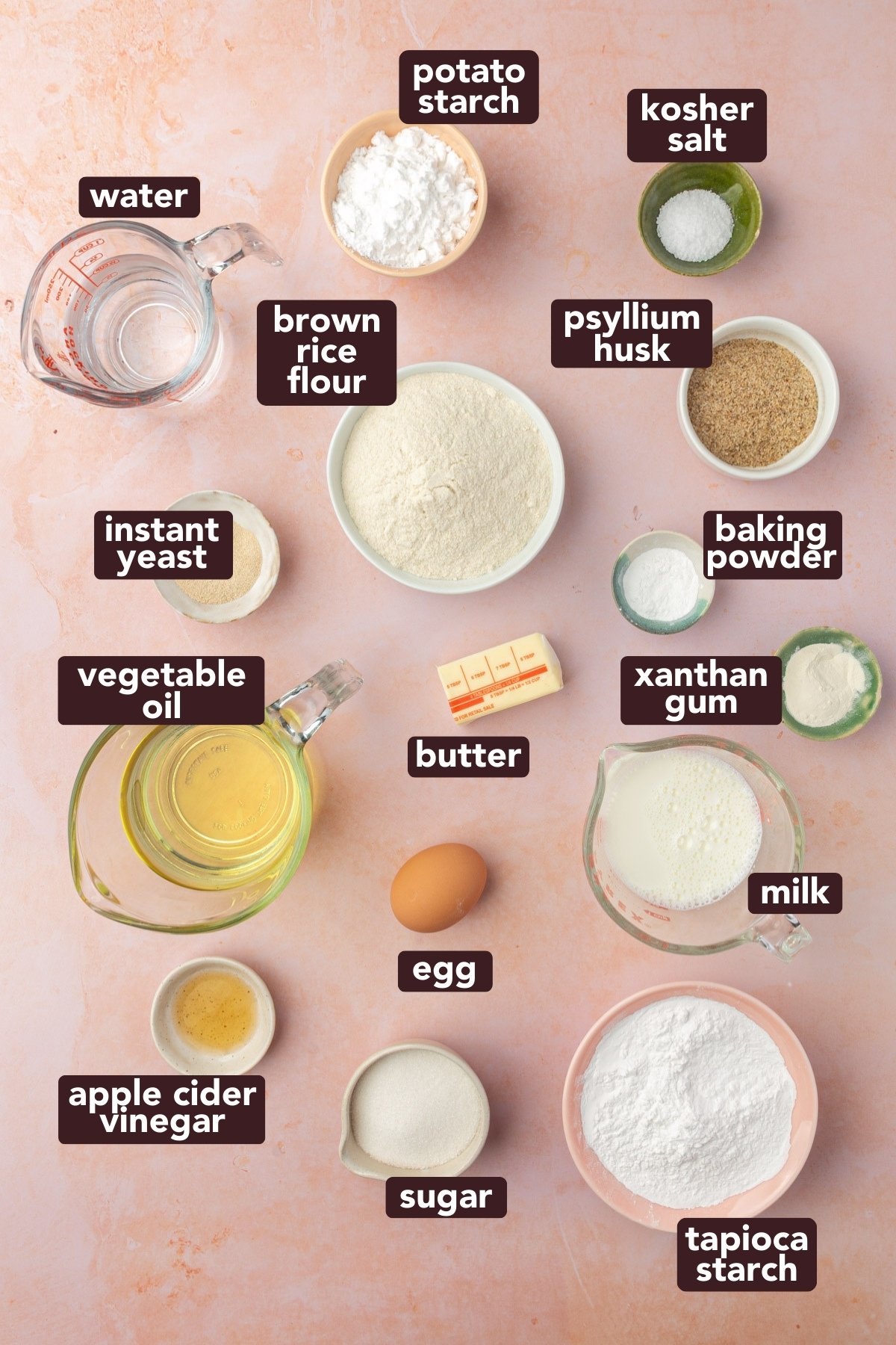 A flat lay of labeled baking ingredients on a pink surface, including potato starch, kosher salt, psyllium husk, baking powder, xanthan gum, milk, tapioca starch, sugar, egg, vinegar, oil, instant yeast, brown rice flour, water, and butter.