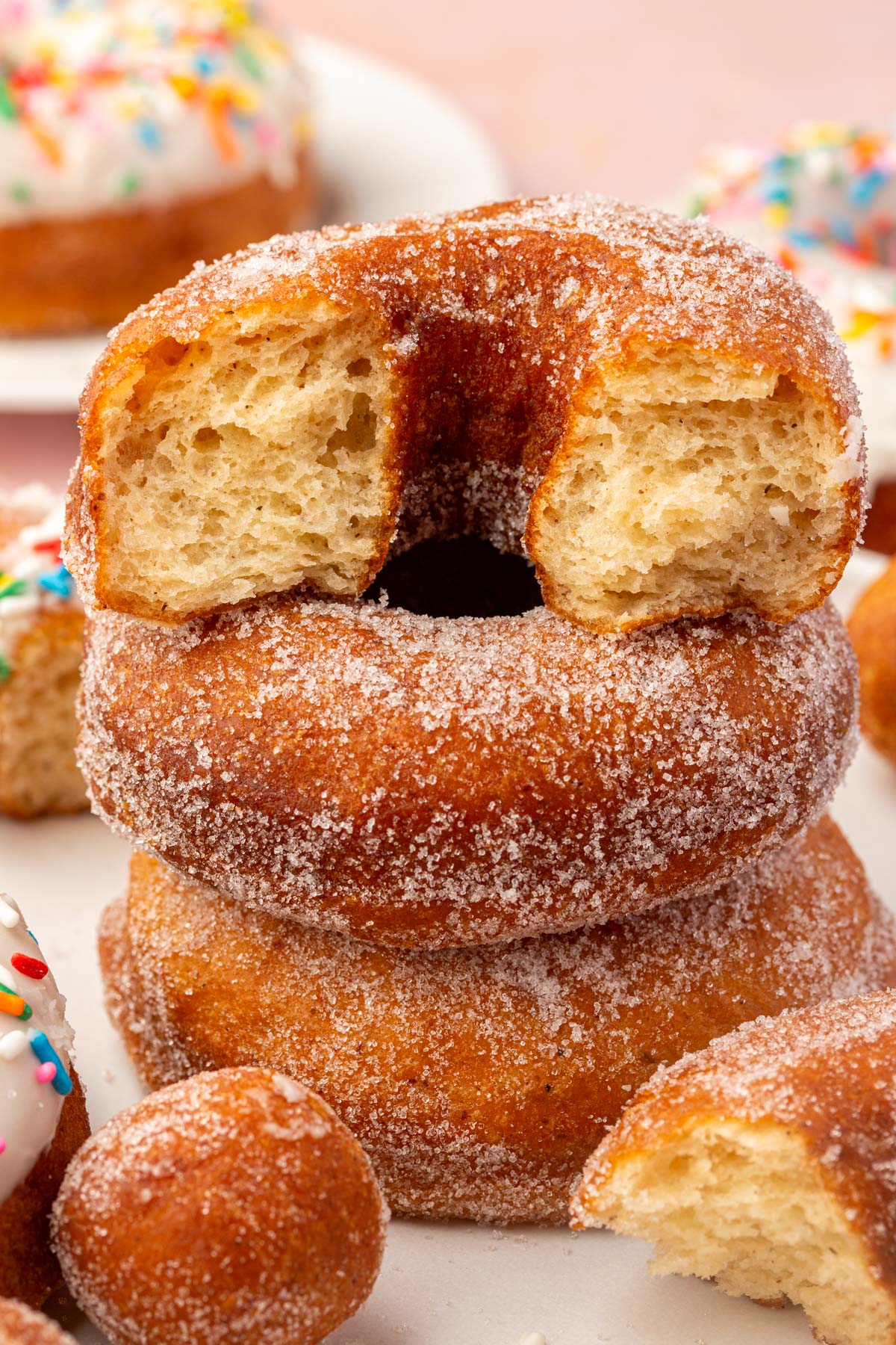 A stack of three sugar-coated donuts, with the top donut missing a bite and showing a fluffy interior. Colorful sprinkled donuts and donut holes are in the background.