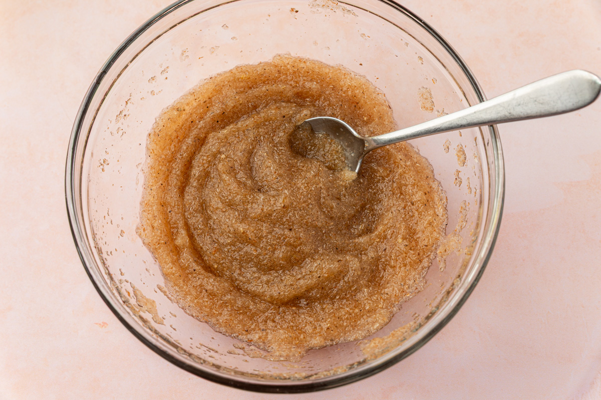 A glass bowl filled with a grainy, light brown sugar scrub mixture, with a metal spoon resting inside, placed on a light pink surface.